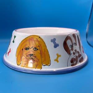 May include: White ceramic dog bowl with a blue rim and a colorful illustration of three dogs and bones. The dogs are a brown and white dog, a yellow dog, and a brown dog.