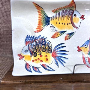 Puede incluir: Plato rectangular de cer&aacute;mica con un dise&ntilde;o pintado a mano de peces coloridos. Los peces tienen detalles amarillos, naranjas, azules y rojos. El plato es blanco con una textura moteada y est&aacute; sobre una superficie de madera.