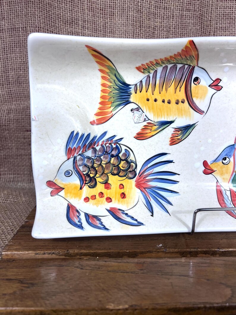 Puede incluir: Plato rectangular de cer&aacute;mica con un dise&ntilde;o pintado a mano de peces coloridos. Los peces tienen detalles amarillos, naranjas, azules y rojos. El plato es blanco con una textura moteada y est&aacute; sobre una superficie de madera.