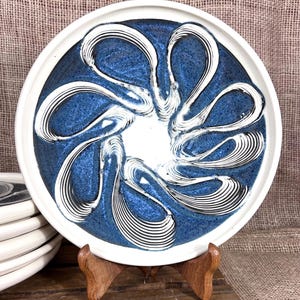 May include: A ceramic plate with a white rim and a blue center featuring a white, swirling design. The plate is displayed on a wooden stand, with a stack of similar plates visible in the background. The plate's diameter is approximately 10 inches.