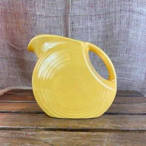 May include: A bright yellow ceramic pitcher with a curved handle and a spout. The pitcher has a smooth, glossy finish and a series of concentric ridges around the body. It sits on a wooden surface.
