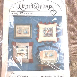 May include: A HeartStrings Country Pleasures pattern booklet in a clear plastic sleeve. The cover features four cross-stitch designs: a framed piece, a ruffled pillow, a square pillow with dark borders, and a small basket with a lid and ribbon.