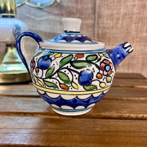 May include: A small, hand-painted ceramic teapot with a lid. The teapot features a blue handle and spout, with a white, blue, yellow, and green floral design. The teapot is sitting on a wooden surface.
