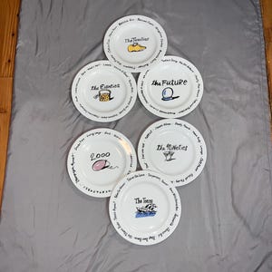 May include: Set of six white ceramic plates with black lettering and illustrations. Each plate features a different decade from the 1920s to the 2000s. The plates are decorated with images representing each decade, such as a pair of shoes for the 1920s, a boombox for the 1980s, and a computer mouse for the 2000s.