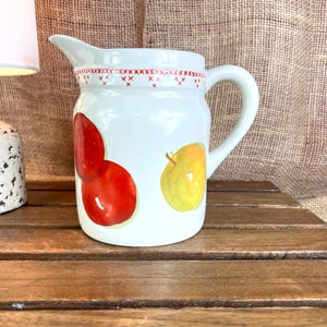 May include: White ceramic pitcher with a red cross-stitch design around the rim. The pitcher features illustrations of a red apple and a yellow apple. The pitcher has a handle and a spout.