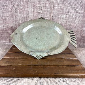 May include: A speckled, light green ceramic fish-shaped platter. The platter has a rounded oval center and a fish tail and head design. Black lines detail the tail and a black dot represents the eye. The platter is displayed on a wooden stand.