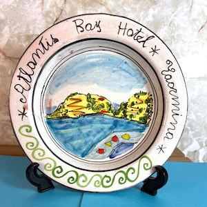 May include: A white ceramic plate with a hand-painted scene of a bay with two islands and a green border. The plate has the words "Bay Hotel * Kalamitsi *" written around the edge.