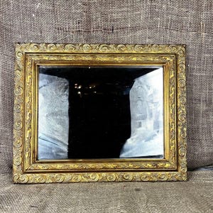 May include: A rectangular wall mirror featuring an ornate gold frame with intricate scrollwork and leaf patterns. The mirror has a reflective glass surface with a dark center and a decorative border that surrounds the entire rectangular edge.