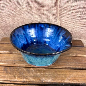 May include: A handmade ceramic bowl with a vibrant blue glaze. The bowl features a dark blue rim and a turquoise base, creating a striking contrast. The interior showcases a flowing, textured pattern. The bowl is set on a wooden surface.