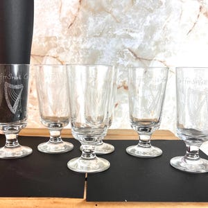 May include: Five clear glass goblets with a harp design etched on the front. The goblets have a round base and a stem. The goblets are all filled with liquid. The goblets are on a black surface.