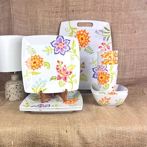 May include: White square dinnerware set with a floral design in orange, purple, and red. The set includes plates, bowls, and a serving tray. The floral pattern is repeated on the cups and bowls, creating a cohesive look.