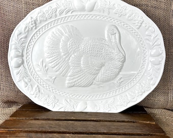 May include: An oval white ceramic platter featuring a raised relief design of a turkey in the center. The rim is decorated with an embossed pattern of various fruits, leaves, and flowers, surrounded by a textured cross-hatch border detail.