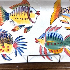 Puede incluir: Una fuente rectangular de cer&aacute;mica con peces pintados a mano en colores vibrantes. Los peces est&aacute;n pintados en tonos amarillos, azules, rojos y naranjas sobre un fondo blanco. La fuente est&aacute; sostenida por un soporte met&aacute;lico, mostrando un dise&ntilde;o decorativo y funcional.