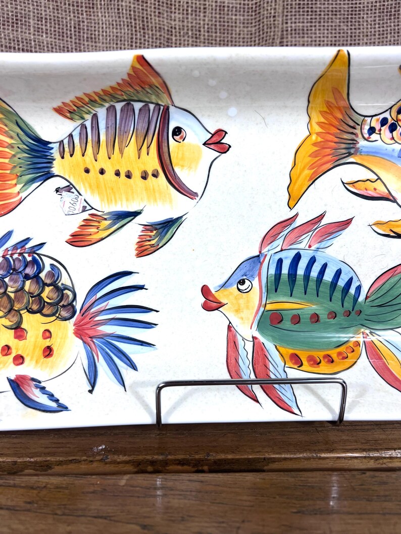 Puede incluir: Una fuente rectangular de cer&aacute;mica con peces pintados a mano en colores vibrantes. Los peces est&aacute;n pintados en tonos amarillos, azules, rojos y naranjas sobre un fondo blanco. La fuente est&aacute; sostenida por un soporte met&aacute;lico, mostrando un dise&ntilde;o decorativo y funcional.