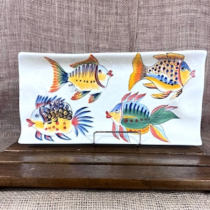May include: Rectangular ceramic serving dish with four colorful fish illustrations. Each fish features a unique design with yellow, orange, blue, and green hues. The dish is supported by a metal stand and rests on a wooden surface.
