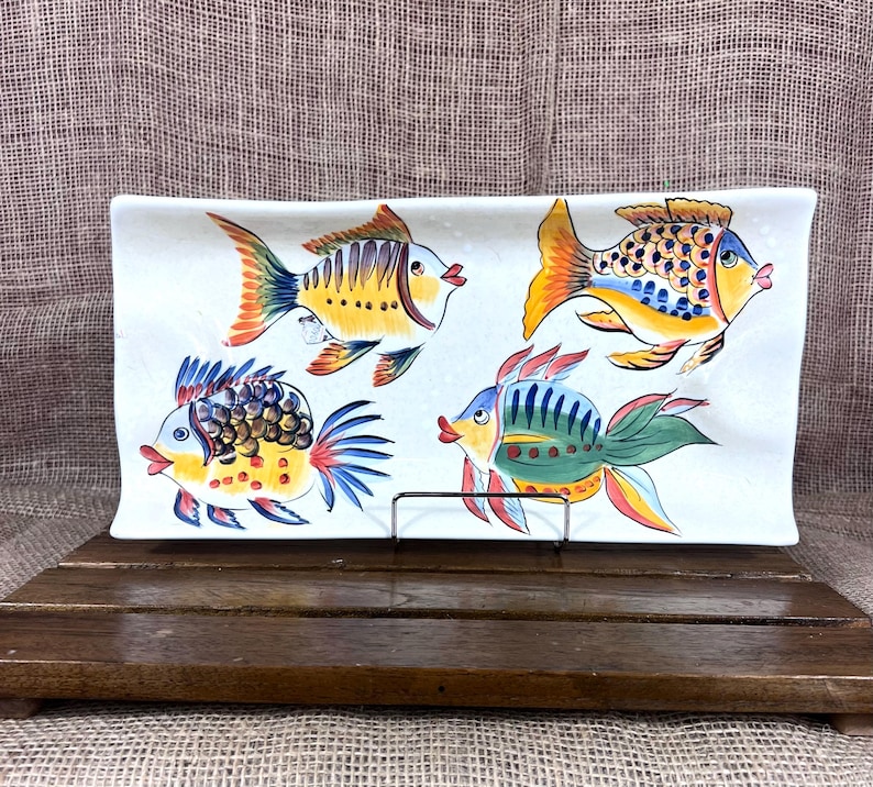 Puede incluir: Plato rectangular de cer&aacute;mica con cuatro ilustraciones de peces coloridos. Cada pez presenta un dise&ntilde;o &uacute;nico con tonos amarillos, naranjas, azules y verdes. El plato est&aacute; sostenido por un soporte met&aacute;lico y descansa sobre una superficie de madera.