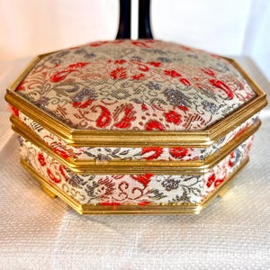 May include: Octagonal shaped trinket box with a gold trim and a floral patterned fabric lining. The fabric is white with red and silver accents.