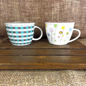 May include: Two ceramic mugs with lids. The left mug features a teal and black buffalo plaid pattern. The right mug is white and decorated with a colorful pattern of stylized flowers on thin stems in shades of green, blue, red, and yellow.