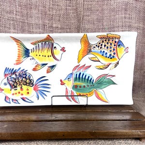 Puede incluir: Una fuente rectangular de cer&aacute;mica blanca con cuatro peces pintados a mano en colores vibrantes. Los peces tienen detalles amarillos, naranjas, azules y verdes. La fuente se exhibe sobre un soporte de madera sobre un fondo neutro.