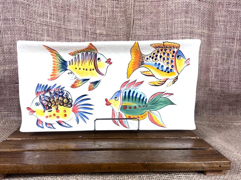 Puede incluir: Una fuente rectangular de cer&aacute;mica blanca con cuatro peces pintados a mano en colores vibrantes. Los peces tienen detalles amarillos, naranjas, azules y verdes. La fuente se exhibe sobre un soporte de madera sobre un fondo neutro.