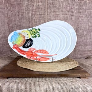 May include: White ceramic seashell-shaped serving platter with a raised design. The platter is decorated with colorful images of seafood, including a red lobster, a blue and yellow scallop, and a black mussel. A lemon slice and parsley are also included.