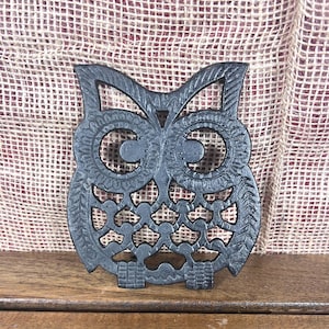 May include: A black cast iron owl-shaped trivet with intricate cut-out details. The owl has large, detailed eyes and a textured surface. The trivet is designed to protect surfaces from hot cookware and adds a decorative touch to the kitchen.