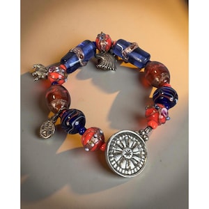 May include: A beaded bracelet featuring a variety of colorful glass beads in shades of blue, red, and brown. The bracelet includes silver charms, including a paw print, a decorative flower, and a stylized eagle. The bracelet is approximately 7 inches in circumference.