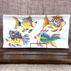 Puede incluir: Plato rectangular de cer&aacute;mica blanca con cuatro ilustraciones de peces coloridos. Los peces presentan detalles amarillos, naranjas, azules y verdes. El plato se exhibe sobre un soporte de madera.