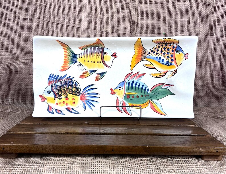 Puede incluir: Plato rectangular de cer&aacute;mica blanca con cuatro ilustraciones de peces coloridos. Los peces presentan detalles amarillos, naranjas, azules y verdes. El plato se exhibe sobre un soporte de madera.