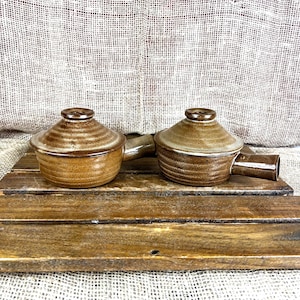 May include: Two matching brown ceramic pots with lids and side handles. Each pot features a rounded base with horizontal ridges and a smooth, conical lid. The glaze is a mottled, earthy brown tone with lighter highlights on the lid surfaces.