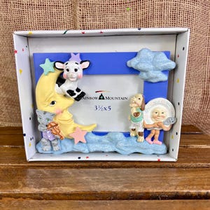 May include: A whimsical photo frame featuring a blue border and a variety of charming, cartoonish characters. The frame includes a cow, a cat playing a violin, a dog, and a figure holding a plate. The frame is 3.5 x 5 inches.
