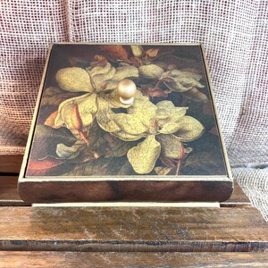 May include: A square wooden box with a floral design on the lid. The lid features a vintage-style painting of yellow and brown flowers. The box has a small, round, gold-colored knob and sits on a wooden surface.
