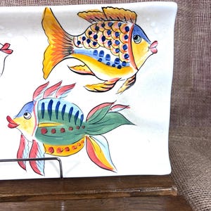 Puede incluir: Una fuente rectangular de cer&aacute;mica blanca con dise&ntilde;os de peces coloridos pintados a mano. Los peces presentan tonos amarillos, azules, verdes y rojos. La fuente se exhibe sobre una superficie de madera, mostrando un tema n&aacute;utico o costero.
