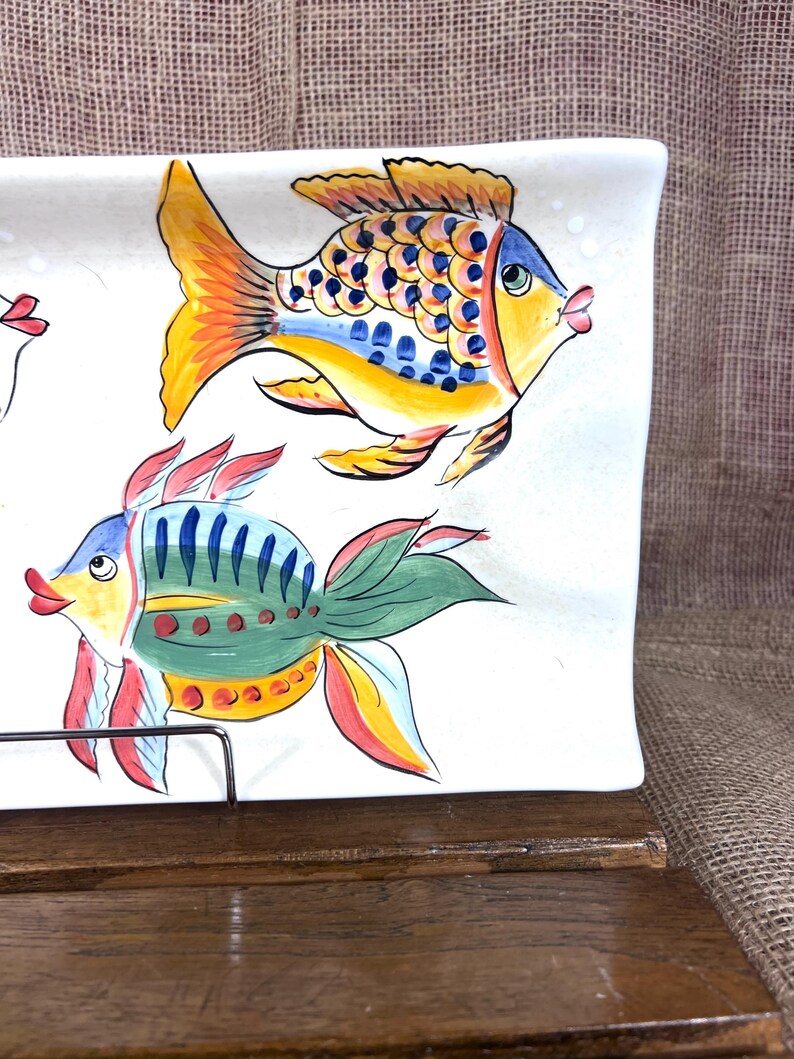 Puede incluir: Una fuente rectangular de cer&aacute;mica blanca con dise&ntilde;os de peces coloridos pintados a mano. Los peces presentan tonos amarillos, azules, verdes y rojos. La fuente se exhibe sobre una superficie de madera, mostrando un tema n&aacute;utico o costero.