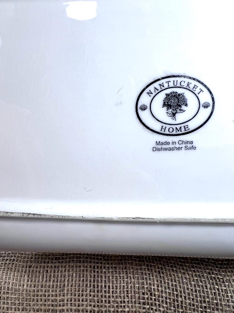 Puede incluir: Una fuente rectangular de cer&aacute;mica blanca con un logotipo ovalado negro que dice "Nantucket Home". El logotipo presenta un &aacute;rbol y las palabras "Made in China Dishwasher Safe".