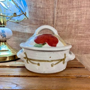 May include: A small, white ceramic lidded dish with a red floral design on the lid. The dish has two small handles and a brown decorative pattern around the body. The lid has a loop handle.