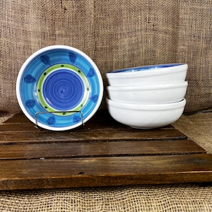May include: A set of four ceramic bowls. One bowl is displayed upright, showing a blue and green hand-painted pattern with a central blue circle, a green ring, and blue leaf-like shapes. The other three bowls are stacked, showing plain white exteriors.