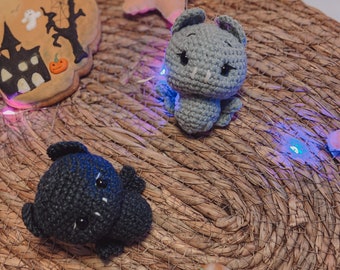 May include: Two crocheted black bats and one gray crocheted bat on a woven brown surface with a gingerbread cookie shaped like a house and a gingerbread cookie shaped like a pumpkin.