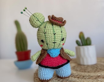 May include: A crocheted cactus toy wearing a brown cowboy hat, a red vest, and blue jeans. The cactus has a green body with a smiling face and is standing on a woven mat.