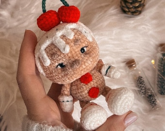 May include: A handmade crochet gingerbread man doll with red cherry accents on top. The doll has white icing details, red button accents, and white feet. The doll is held in a hand against a soft, white background.