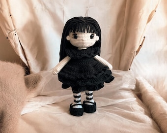 Könnte beinhalten: Eine gehäkelte Puppe mit schwarzen Haaren, einem schwarzen Kleid mit Rüschen und schwarz-weiß gestreiften Strümpfen. Die Puppe trägt schwarze Schuhe.