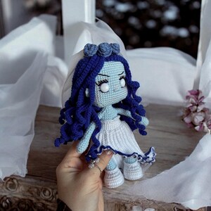 May include: A crocheted doll with blue hair, white eyes, and a white dress. The doll is wearing a veil and has a blue and white trim on the dress.