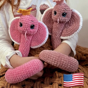 Può includere: Due giocattoli a forma di serpente in crochet rosa con bordi bianchi e corone gialle. I serpenti sono arrotolati su una coperta marrone.