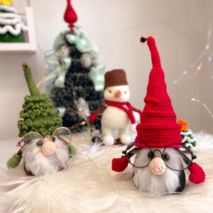 Könnte beinhalten: Zwei gehäkelte Gnome mit Brille, einer mit grünem Hut und der andere mit rotem Hut, sitzen auf einer weißen, flauschigen Oberfläche. Im Hintergrund sind ein Schneemann und ein kleiner Weihnachtsbaum zu sehen.