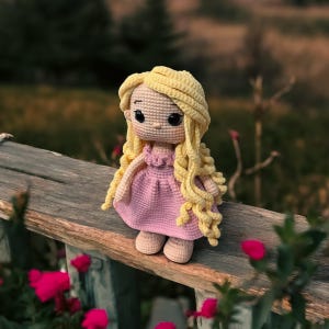 May include: A crocheted doll with long blonde hair wearing a pink dress. The doll is standing on a wooden surface.