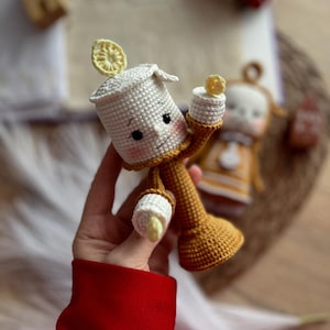 May include: A crocheted plush toy of Lumiere, the candelabra from Disney's Beauty and the Beast. The toy is white and yellow with a brown base and has a friendly expression.
