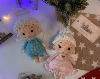 May include: Two crocheted angel dolls with white wings. The angel on the left is wearing a light blue dress and the angel on the right is wearing a pink dress. Both dolls have white yarn hair and embroidered facial features.
