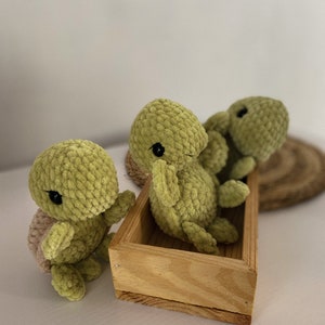 Puede incluir: Tres peluches de tortuga de crochet verde, dos en una caja de madera y uno en la mesa. Las tortues tienen ojos negros y un caparazón marrón.