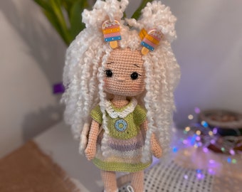 Puede incluir: Una muñeca de crochet con pelo blanco rizado, que lleva un vestido verde y morado con un diseño floral. La muñeca tiene zapatos blancos y sostiene dos pinzas para el pelo en forma de paleta de colores.