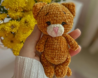 May include: A small, crocheted plush toy in the shape of a brown cat with a white face and pink nose. The cat is being held by a hand wearing a white sweater.